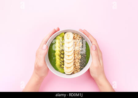 Woman's hands holding smoothie aux épinards avec bol de granola, quinoa, banane et kiwi sur fond rose. Vue supérieure de style minimal. Banque D'Images