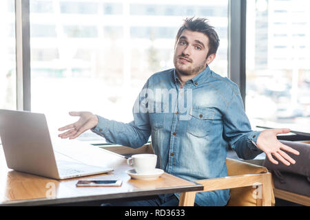 Pas sûr ! Portrait de confondre beau jeune barbu freelancer dans blue jeans chemise sont assis dans un café et travailler sur ordinateur portable avec les bras levés et pu Banque D'Images