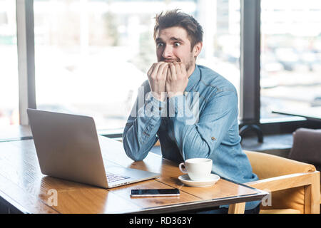 Peur ! Vue de côté portrait de jeune homme nerveux émotionnel en bleu jeans chemise sont assis dans un café, travaillant lui-même se ronger les ongles et onlone caus Banque D'Images