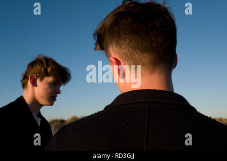 Deux adolescents à la cool, l'un avec l'arrière de la tête et des épaules en premier plan, se tenant ensemble sur une plage près de la mer au coucher du soleil en France Banque D'Images