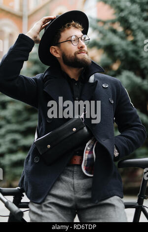 Portrait de beau barbu dans les tenues à l'écart et souriant tout en se penchant sur son vélo standing outdoors Banque D'Images