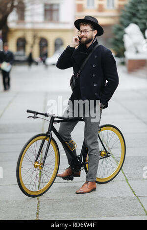 Heureux jeune homme barbu en conversation sur le téléphone mobile et souriant alors qu'il était assis près de son vélo en plein air Banque D'Images