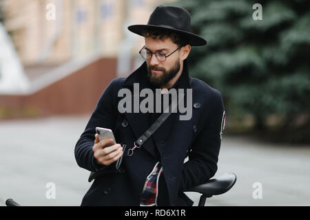 L'insouciance du temps dans la ville. Low angle view of young bearded man holding mobile phone et à le regarder alors qu'il était assis à l'extérieur Banque D'Images