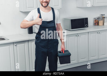 Vue partielle de l'ensemble de travail réparateur à barbu qui maintient la boîte à outils et showing thumb up dans la cuisine à la maison Banque D'Images