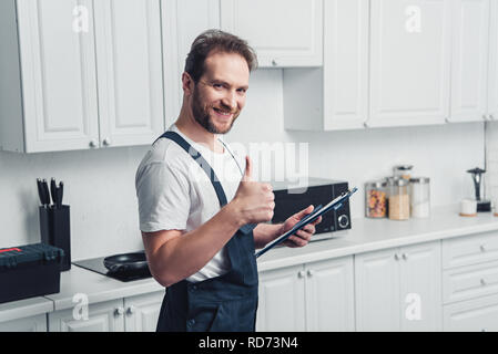 Handsome smiling repairman barbus adultes holding clipboard et faisant thumb up geste en cuisine Banque D'Images