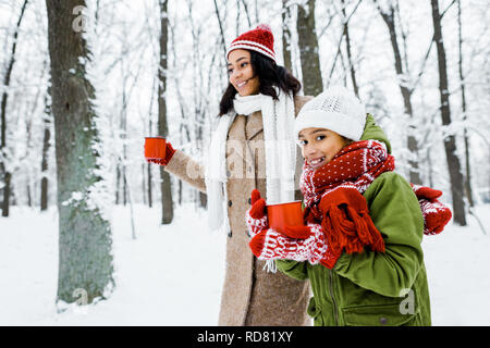 Belle mère afro-américaine et mignon fille walking and holding red cups in snowy forest Banque D'Images