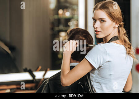 Belle jeune hairstylist looking at camera tout en peignant et la coupe de cheveux de clients masculins en instituts de beauté Banque D'Images