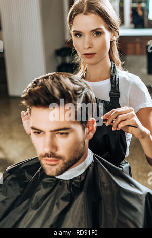 Hairstylist looking at camera tandis que la coupe de cheveux de beau jeune homme en instituts de beauté Banque D'Images