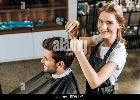 High angle view of hairstylist smiling at camera tandis que la coupe de cheveux de beau client en instituts de beauté Banque D'Images