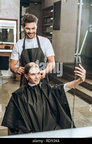 Smiling girl en tenant avec selfies smartphone pendant que hairstylist faisant en salon de coiffure Banque D'Images