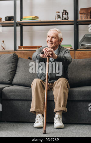 Cheerful pensionné smiling, bâton de marche et sitting on sofa Banque D'Images