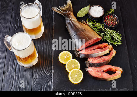 Couper en darnes de saumon sauvage avec du romarin frais sel et poivre de pierre noire sur un plateau avec la mousse de la bière dans des tasses en verre, vue de dessus, close-up Banque D'Images
