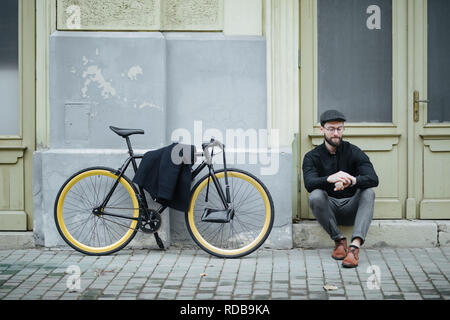 Beau jeune homme assis sur le hipster road et posant avec sac à dos et vélo sur le coucher du soleil Banque D'Images