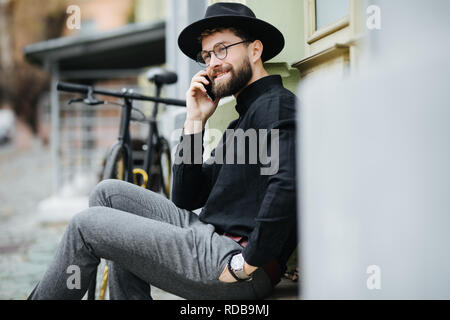 Invite des amis pour un tour. Heureux jeune homme barbu en conversation sur le téléphone mobile et souriant alors qu'il était assis près de son vélo en plein air Banque D'Images