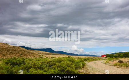 Chemin de terre bordé par des prairies arides et les armoises avec grange rouge sur l'horizon et les Rocheuses en été dans la région de Cody, Wyoming, USA. Banque D'Images