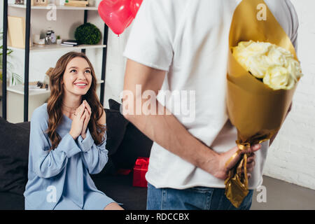 Young man holding bouquet de roses derrière le dos en surpris smiling girl sitting avec les mains pliées sur canapé Banque D'Images