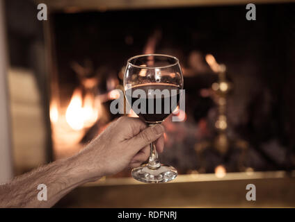 Close up of man holding et de boire un verre de vin rouge par cheminée bois ouvert se détendre et profiter de moment de calme à la maison en hiver maison de vacances, Chr Banque D'Images