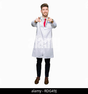 Beau jeune homme portant un manteau médical médecin pointant vers vous et l'appareil photo avec les doigts, souriant et de bonne humeur positive Banque D'Images