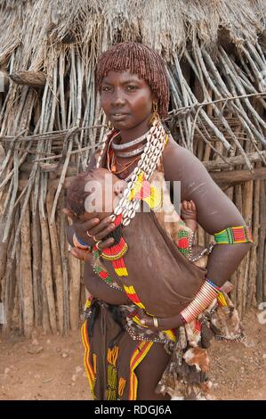 Jeune femme Hamar avec coiffure traditionnelle et d'argile rouge colliers faits de cauris et tenant son bébé dans les bras Banque D'Images