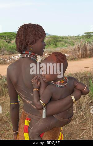 Jeune femme Hamar avec coiffure traditionnelle en argile rouge tenant un bébé dans ses bras et avec les cicatrices et les marques de coups de fouet Banque D'Images