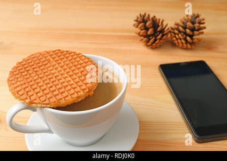 Café chaud avec Stroopwafel placé sur le dessus de la Tasse Servi sur table en bois avec Smart Phone floue et pommes de pin sec en arrière-plan Banque D'Images