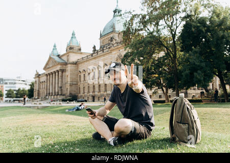 Jeune homme étudiant ou touristique assis sur l'herbe dans les rues de Leipzig en Allemagne et montre le geste de la main signifie la paix. Banque D'Images