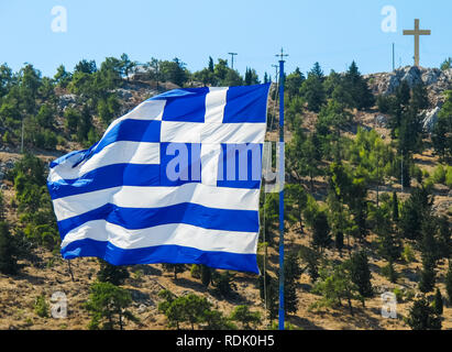 Le drapeau national de la Grèce sur le mât se développe dans le vent contre l'arrière-plan d'une colline avec des arbres verts et une croix sur le dessus. Île grecque de Banque D'Images