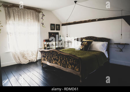 Vue intérieure d'une chambre mansardée avec parquet en bois sombre et des murs blancs, un lit double français avec jet vert et dentelle rideau à la fenêtre. Banque D'Images