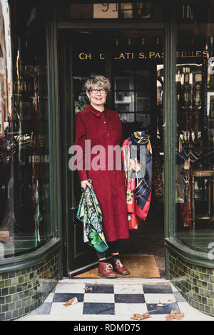 Smiling senior woman wearing glasses et robe rouge debout avant de interior design store, looking at camera. Banque D'Images