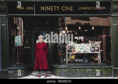 Smiling senior woman wearing glasses et robe rouge debout avant de interior design store, looking at camera. Banque D'Images