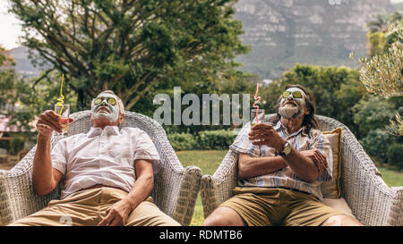 Les hommes âgés avec masque facial et tranches de concombre sur le visage au spa. Amis retraités bénéficiant de soins spa du visage avec des lunettes de jus à la main. Banque D'Images