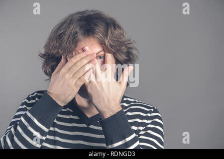 Bel homme avec des cheveux bouclés, de soies, se cache le visage avec les mains, se profile au doigts, porte pullover rayé, timide, se sent plus isolé gray background Banque D'Images