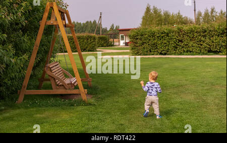 Petit enfant courir vers balançoire en bois banc dans le parc. Enfant jouant dans le jardin. Banque D'Images