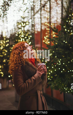 Belle jeune fille rousse holding paper verre avec verre. Portrait de nuit rue de la ville. Bokeh de lumières de fête. Banque D'Images