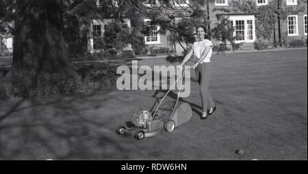 1950s, historique, une élégante dame portant des pantalons tenant les poignées d'une nouvelle tondeuse à moteur, avec une lame rotative, à l'extérieur sur la pelouse d'un grand jardin de maison de campagne, Angleterre, Royaume-Uni. Banque D'Images