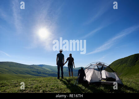 Paysage incroyable des Carpates sous le ciel bleu avec un soleil d'été. Vue arrière d'un jeune couple appréciant les mains près de la tente, sac à dos et bâtons de randonnée Banque D'Images