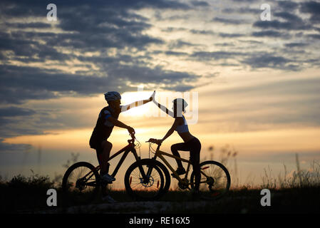 Couple adorable dans la prise casque en famille sur les bicyclettes, debout et poser. Silhouettes d'homme et femme highing sportive cinq. Incroyable coucher du soleil et ciel magnifique arrière-plan. Banque D'Images