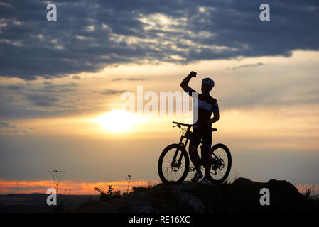 Smiling man sportive est dotée d'un vélo sur la roche au sommet d'une colline sur un arrière-plan flou d'un beau coucher du soleil. L'homme est vêtu de vêtements de sport et un casque Banque D'Images