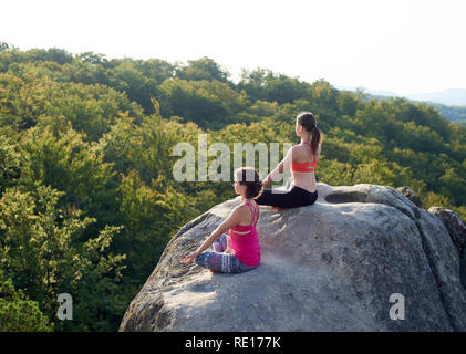 Vue du haut de deux pieds nus athlétiques women sitting in lotus pose sur le dessus de l'énormes rochers sur le vert des montagnes et le fond de ciel bleu clair. Le yoga, fitness et d'un style de vie sain concept. Siddhasana Banque D'Images