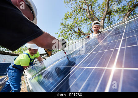 Équipe de trois techniciens qui travaillent à l'extérieur de l'installation du système de panneaux solaires photovoltaïques en milieu rural sur la campagne lumineuse, ensoleillée journée d'été. Les énergies renouvelables de production d'énergie verte bon marché écologique concept. Banque D'Images