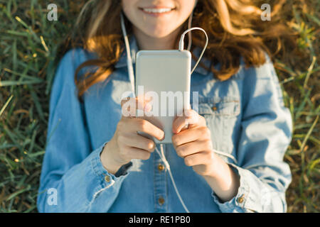 Teenage girl in hat avec des écouteurs se trouve sur l'herbe verte et ressemble au téléphone. L'accent sur smartphone, vue d'en haut. Banque D'Images