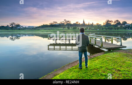 Homme debout sur un petit pont reflétant sur le lac au lever du soleil comme une façon d'accueillir la belle nouvelle journée Banque D'Images