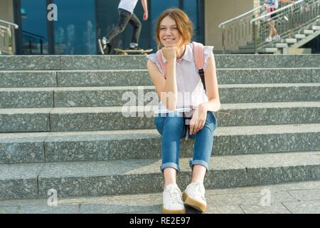 Portrait plein air de 14 ans lycéenne. Jeune fille avec sac à dos, s'assoit sur les marches de l'école détient dans ses mains de l'ordinateur portable Banque D'Images