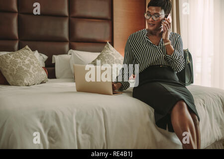 African businesswoman dans l'hôtel, à l'aide de l'ordinateur portable et le smartphone assis sur le lit. Femelle adulte en chef de la chambre d'hôtel moderne assis sur le lit et faisant affaires Banque D'Images