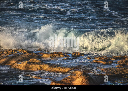 Avec des vagues se brisant sur des rochers de pulvérisation sur la rive au coucher du soleil Banque D'Images