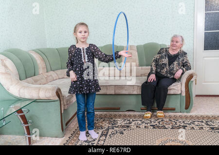 Petite-fille fait tourner le cerceau sur le bras, en face de son arrière grand-mère, assise sur le divan Banque D'Images