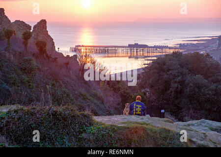 Hastings, Royaume-Uni. 20 janvier 2019. Une soirée ensoleillée calme sur un garçon et son chien s'asseoir et regarder le coucher de soleil à côté de la ruine du château, donnant sur le quai et la mer au loin Beachy Head. Banque D'Images