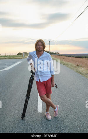 Le beau jeune homme se brutal sur la route au coucher du soleil, il est habillé d'une chemise blanche avec un manche courte et short rouge, le photographe Banque D'Images