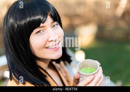 Happy smiling Woman holding tea cup face closeup en extérieur dans le jardin avec fille et vert matcha cheveux noir Banque D'Images