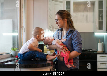 Jeune mère détient sa petite fille avec sucette. Fils aîné est assise pieds nus sur le tableau dans la cuisine et la holding thermomètre électronique. Il l'embrasse Banque D'Images